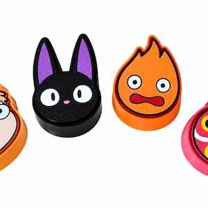 May include: Four circular decorative charms featuring colorful character designs. From left to right: a light-skinned face with orange hair, a black cat head with purple ears, an orange flame character with wide eyes, and a pink mask with yellow eyes.
