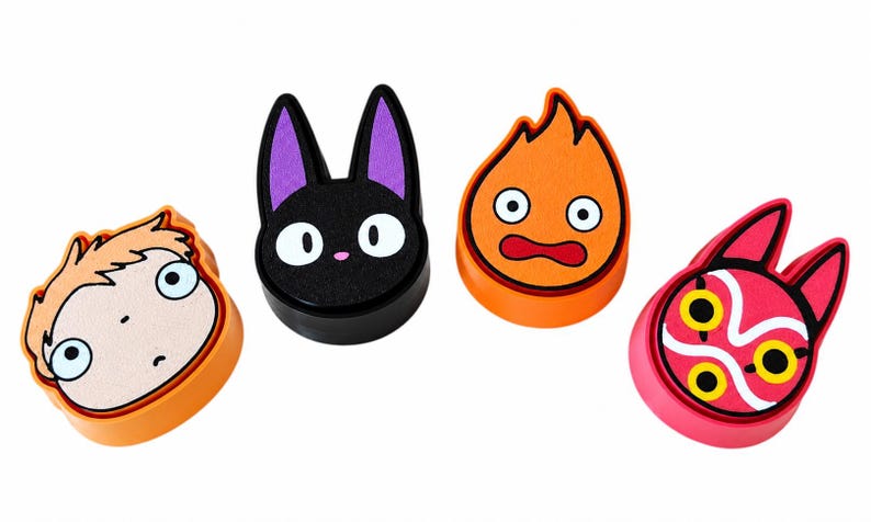 May include: Four circular decorative charms featuring colorful character designs. From left to right: a light-skinned face with orange hair, a black cat head with purple ears, an orange flame character with wide eyes, and a pink mask with yellow eyes.