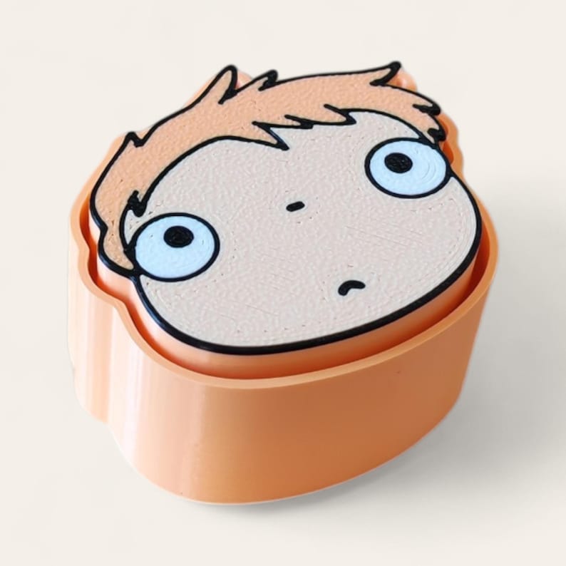May include: A peach-colored plastic cookie cutter shaped like a cartoon character head with light brown hair, large white eyes with black pupils, a small nose, and a curved mouth. The object has a smooth, solid base and a raised top edge.