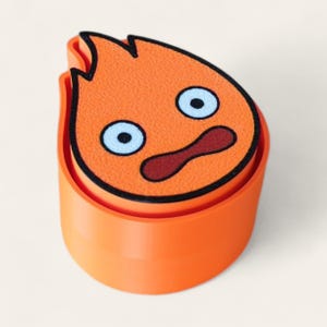 May include: A cylindrical orange container featuring a textured orange flame-shaped lid with a cartoon face. The face has two large light blue eyes with black pupils and a dark red mouth, all outlined in black, set against a solid orange base.