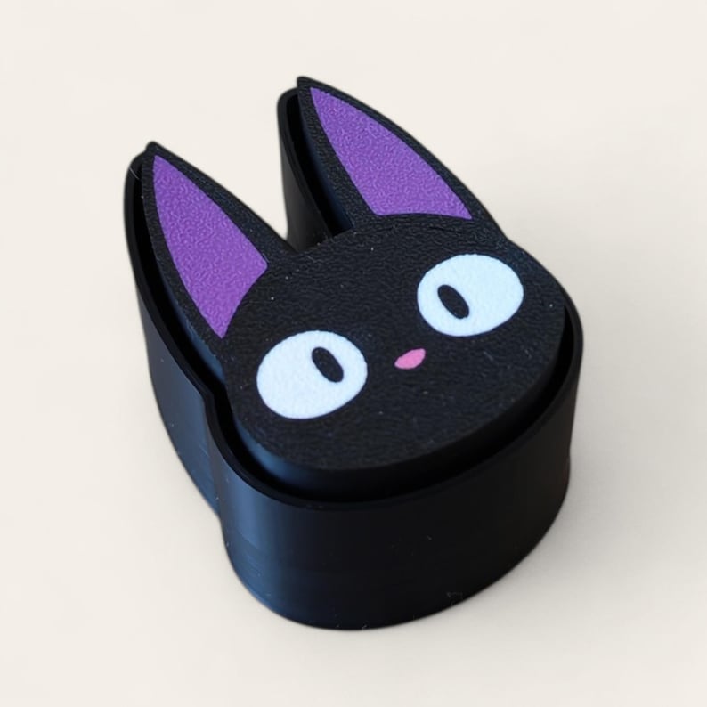 May include: A black cat-shaped object with a flat top surface featuring a stylized black cat face. The cat has large white oval eyes, a small pink nose, and pointed ears with purple inner sections. The base is a solid black curved band.