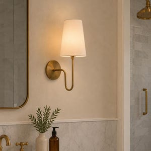 Op de afbeelding: Een messing wandlamp met een witte lampenkap verlicht een badkamer. De lamp heeft een gebogen arm en een cirkelvormige achterplaat. De badkamer heeft een marmeren aanrecht, een spiegel en een douche met messing armaturen.