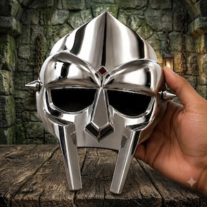 May include: A polished silver-colored metal mask featuring a pointed forehead, two large eye openings, a central nose guard, and two small spikes on the sides. A small, square red accent is positioned at the center of the brow area.