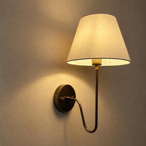 Op de afbeelding: Een wandlamp met een crèmekleurige lampenkap en een messingkleurig metalen frame. De lampenkap heeft een licht taps toelopende vorm en een donkere rand. De lamp is ontworpen voor wandmontage en zorgt voor zachte, sfeervolle verlichting.