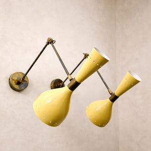 May include: Two yellow wall-mounted lamps with adjustable arms. The lamps have a conical shade with small perforations and a brass base. The arms are dark metal, and the lamps are mounted on a light-colored wall. The lamps are a vintage style.
