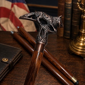 May include: A wooden walking stick featuring a bronze wolf head handle. The dark brown shaft has a brass collar and tip. The background includes an American flag, books, and a leather wallet.