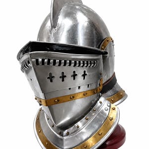 May include: A polished steel knight helmet featuring a rounded dome, a movable visor with horizontal slits and cross-shaped ventilation holes, and brass-colored metal trim accents secured with small rivets along the neck guard and visor base.