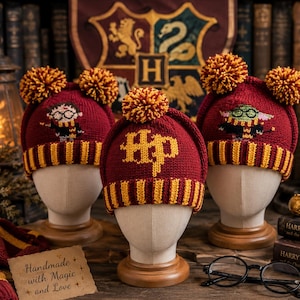 Könnte beinhalten: Drei weinrote und goldfarbene Strickmützen mit Bommeln, jede mit einem anderen Design: Harry Potter, das HP-Logo und eine Figur. Die Mützen werden auf Schaufensterpuppenköpfen ausgestellt. Ein Schal und Bücher sind im Hintergrund.