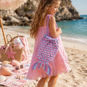 May include: A pink dress and a mermaid-tail-shaped crochet bag. The bag features a drawstring closure and a tail with blue, purple, and pink stripes. The beach scene includes a straw bag, a pink beach umbrella, and seashells.