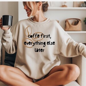 May include: A beige crewneck sweatshirt featuring a graphic of three tan coffee bean outlines and the black text "coffe first, everything else later" printed on the front. The garment has long sleeves and a ribbed collar, cuffs, and hem.
