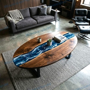 Op de afbeelding: Ovale houten salontafel met een blauwe hars rivier ontwerp. De tafel heeft een donkere houten afwerking en zwarte metalen poten. Een kleine plant en boeken liggen op de tafel.
