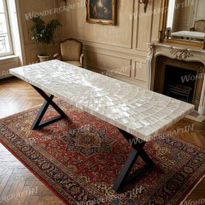 Peut inclure: Une table de salle à manger rectangulaire blanche avec une surface en mosaïque, soutenue par des pieds en métal noir. La table est posée sur un tapis à motifs rouge et marine, avec une cheminée et un miroir en arrière-plan. La table mesure environ 183 cm de long.
