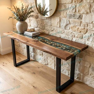 May include: A wooden console table with a river of stones encased in clear resin. The table has a natural wood grain finish and black metal legs. A vase with dried plants and books are on the table.