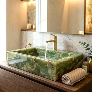 May include: A rectangular green onyx bathroom sink with a gold tap. The sink is made of polished, veined stone. A rolled beige towel and a bottle of lotion sit on the wooden vanity. The mirror reflects the sink and tap.