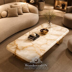 May include: A rectangular coffee table featuring a polished cream and gold veined stone top with rounded corners, supported by a gold metal base. On the surface sits a small gold bowl, a clear glass vase with greenery, and two stacked books.