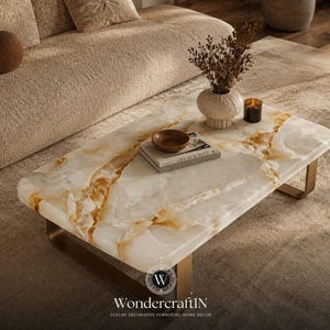 May include: A rectangular coffee table featuring a polished white and gold-veined stone top supported by a metallic base. The table holds a small wooden bowl, a book titled Live Beautiful, a textured white vase with dried foliage, and a candle.