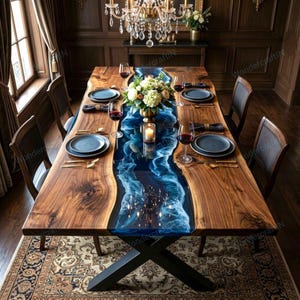 May include: A large wooden dining table with a blue resin river design. The table is set with plates, silverware, and wine glasses. A floral centerpiece and candles adorn the table. The table is supported by a black metal base.