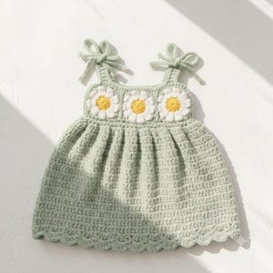 May include: A light sage green crochet dress featuring three white daisy motifs with yellow centers across the chest. The garment has two shoulder straps that tie into bows and a scalloped hemline, crafted with a textured yarn stitch pattern.