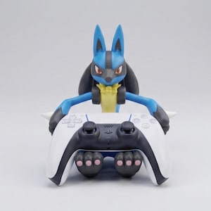 May include: A blue and black Lucario figurine holding a white and black video game controller. The character has pointed ears, red eyes, and a yellow chest tuft. The controller features black analog sticks and buttons. The text Geek & Gaming Studio is visible.