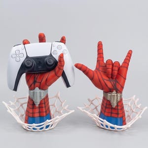 Peut inclure: Deux sculptures de mains de super-héros rouges et bleues émergeant de bases en forme de toile blanche. Une main tient une manette de jeu vidéo blanche et noire, tandis que l'autre est en geste rock. Les poignets portent des lance-toiles métalliques.