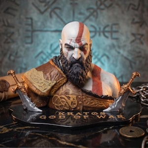 Pode incluir: Um busto detalhado de Kratos de God of War, apresentando uma tez pálida com uma tatuagem vermelha, barba escura e armadura de couro castanho. A figura está montada numa base preta com o texto KRATOS GOD OF WAR e duas lâminas prateadas.