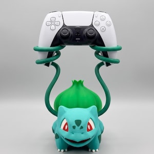 May include: A teal and green creature figurine with a bulbous plant on its back, featuring two long, spiraling green vines extending upward to hold a white and black wireless video game controller. The text Geek & Gaming Studio is visible.