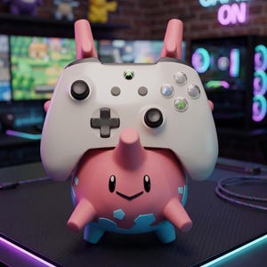 Puede incluir: Un soporte para mando con forma de personaje rosa y azul que sostiene un mando inalámbrico Xbox blanco. El soporte presenta un cuerpo redondeado, extremidades pequeñas y un cuerno central, proporcionando una base estable para el periférico.
