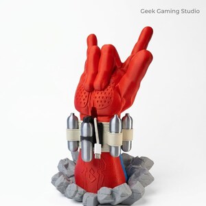 May include: A red hand sculpture in a rock-and-roll gesture emerging from a gray rocky base. The wrist features a beige strap holding four silver canisters and a white USB-C charging cable. The text Geek Gaming Studio is visible.