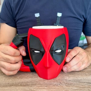 May include: A red and black ceramic mug shaped like the Deadpool mask, featuring large black eye patches with white centres. The handle is designed to look like a black handgun, and two black sword hilts protrude from the top rim of the mug.