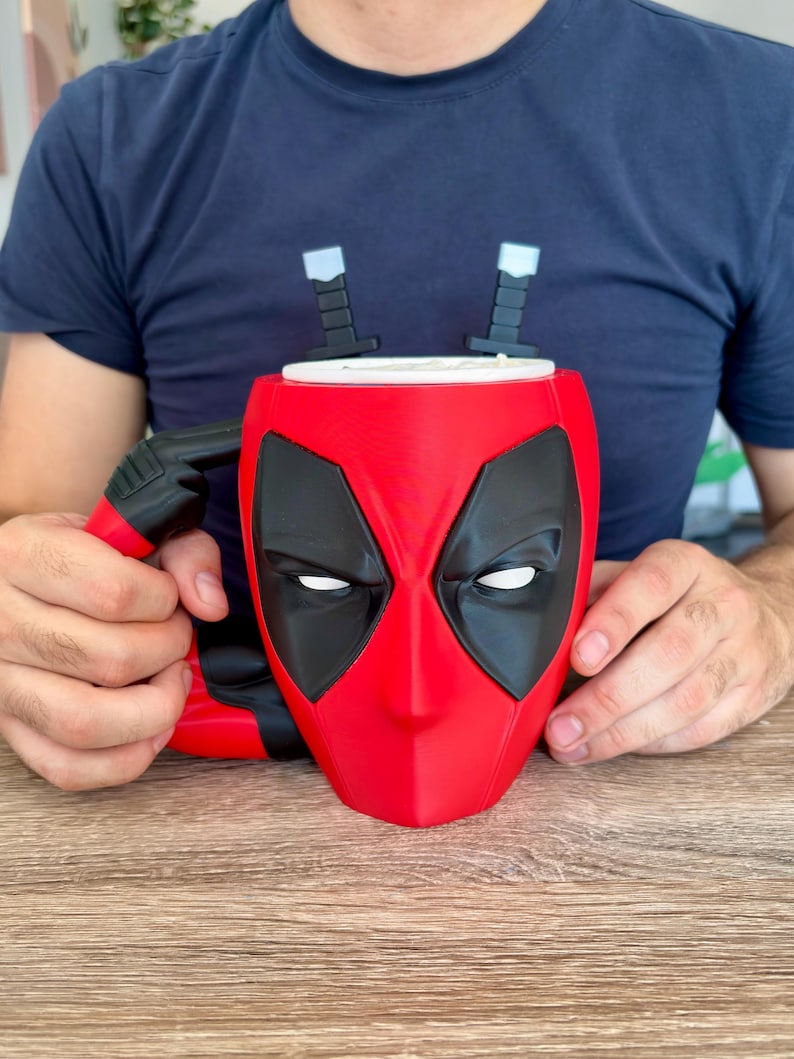 May include: A red and black ceramic mug shaped like the Deadpool mask, featuring large black eye patches with white centres. The handle is designed to look like a black handgun, and two black sword hilts protrude from the top rim of the mug.