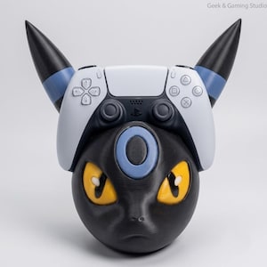 Könnte beinhalten: Ein schwarz-blauer Gaming-Controller-Ständer in Form eines fiktiven Kreaturenkopfes mit gelben Augen und spitzen Ohren. Der Ständer zeigt einen weißen und schwarzen Game-Controller, der darauf ruht, mit dem Text Geek & Gaming Studio.