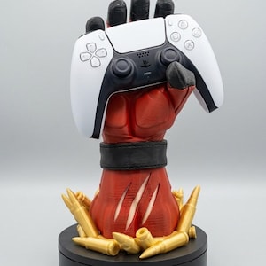 May include: A red, muscular hand sculpture with black fingers and a black wristband, holding a white and black video game controller. The base is surrounded by gold-colored bullet casings, and the forearm features three vertical claw-like slashes.