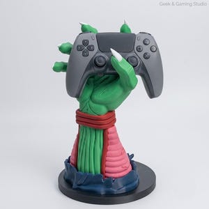 Puede incluir: Una escultura de mano verde con garras blancas que sostiene un mando de videojuego gris. La mano presenta una muñequera roja y una sección de antebrazo rosa texturizada, todo sobre una base negra circular. El texto Geek & Gaming Studio es visible.