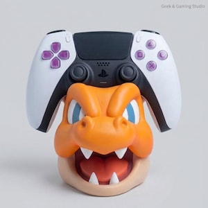 May include: A white and black video game controller resting on an orange and tan character-shaped stand. The stand features a sculpted head with blue eyes, sharp white teeth, and an open mouth. The text Geek & Gaming Studio appears in the corner.
