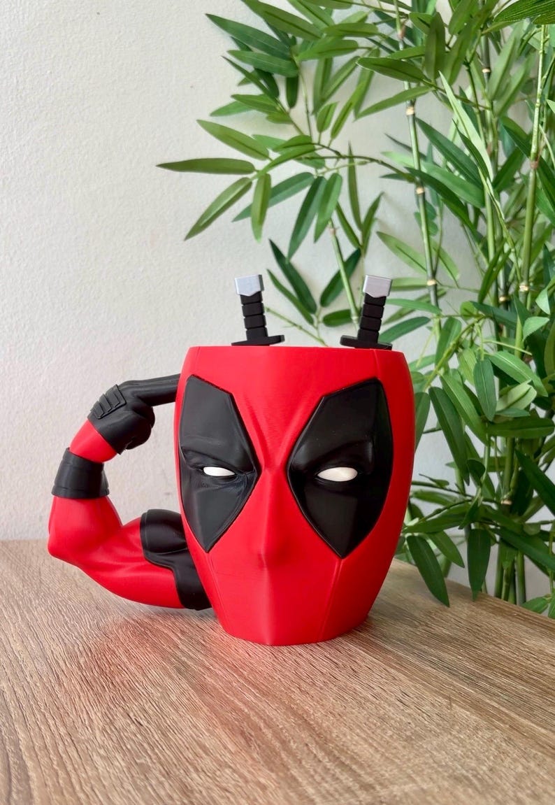 May include: A red and black mug shaped like the head of the character Deadpool, featuring white eye patches and a handle designed as a muscular arm pointing to the head. Two small black and silver sword hilts protrude from the top of the mug.