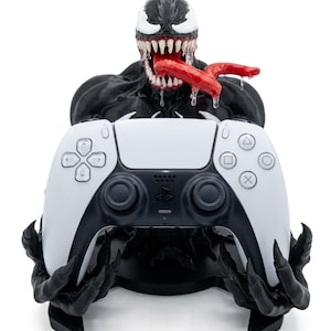 Puede incluir: Una figura negra del personaje Venom que actúa como soporte para mando, con una boca ancha de dientes afilados y una larga lengua roja. La figura sostiene un mando de juego inalámbrico blanco y negro en sus manos, con el mando apoyado.