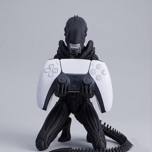 Pode incluir: Uma estatueta preta de Xenomorfo em posição de joelhos, segurando um controle de videogame branco e preto. A criatura tem uma cabeça lisa e alongada, dentes visíveis e uma longa cauda segmentada na base. Texto: Geek & Gaming Studio.