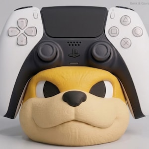 May include: A black and white video game controller resting on a yellow and beige animal head stand. The controller features a directional pad, two analogue sticks, and four action buttons. The stand has a textured surface and a black nose.