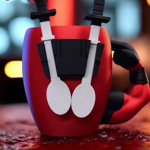 May include: A red ceramic mug featuring a black harness on the front holding two white spoon-shaped utensils with black handles. The mug handle is shaped like a muscular red arm with a black glove, mimicking a character design with a red and black theme.
