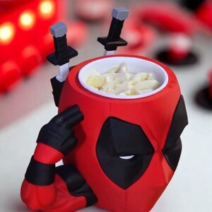 May include: A red and black mug shaped like the head of a character with a mask, featuring two black sword handles protruding from the back and a sculpted arm with a black glove pointing to the temple. The interior of the mug is white.