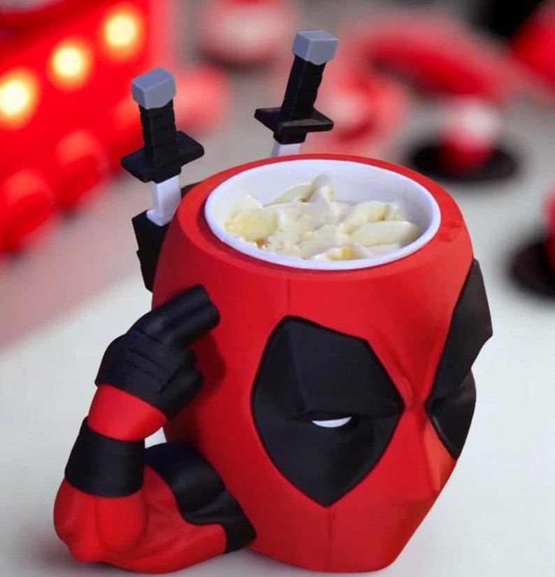 May include: A red and black mug shaped like the head of a character with a mask, featuring two black sword handles protruding from the back and a sculpted arm with a black glove pointing to the temple. The interior of the mug is white.