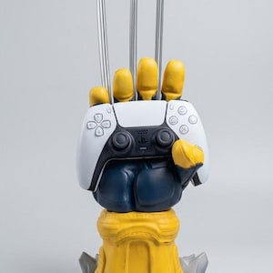May include: A yellow and blue hand-shaped controller stand featuring three long silver claws extending upward. The hand holds a white and black wireless game controller. The base is yellow with grey rock-like accents at the bottom.