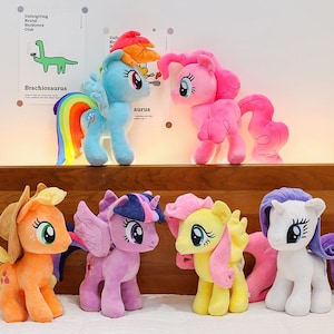 Könnte beinhalten: Eine Sammlung von sechs Plüsch-Pony-Spielzeugen in verschiedenen Farben, darunter hellblau mit einer Regenbogenmähne, leuchtendes Pink, Orange mit Hut, Lila mit Horn, Gelb mit rosa Mähne und Weiß mit violetter Mähne, alle mit großen Augen.