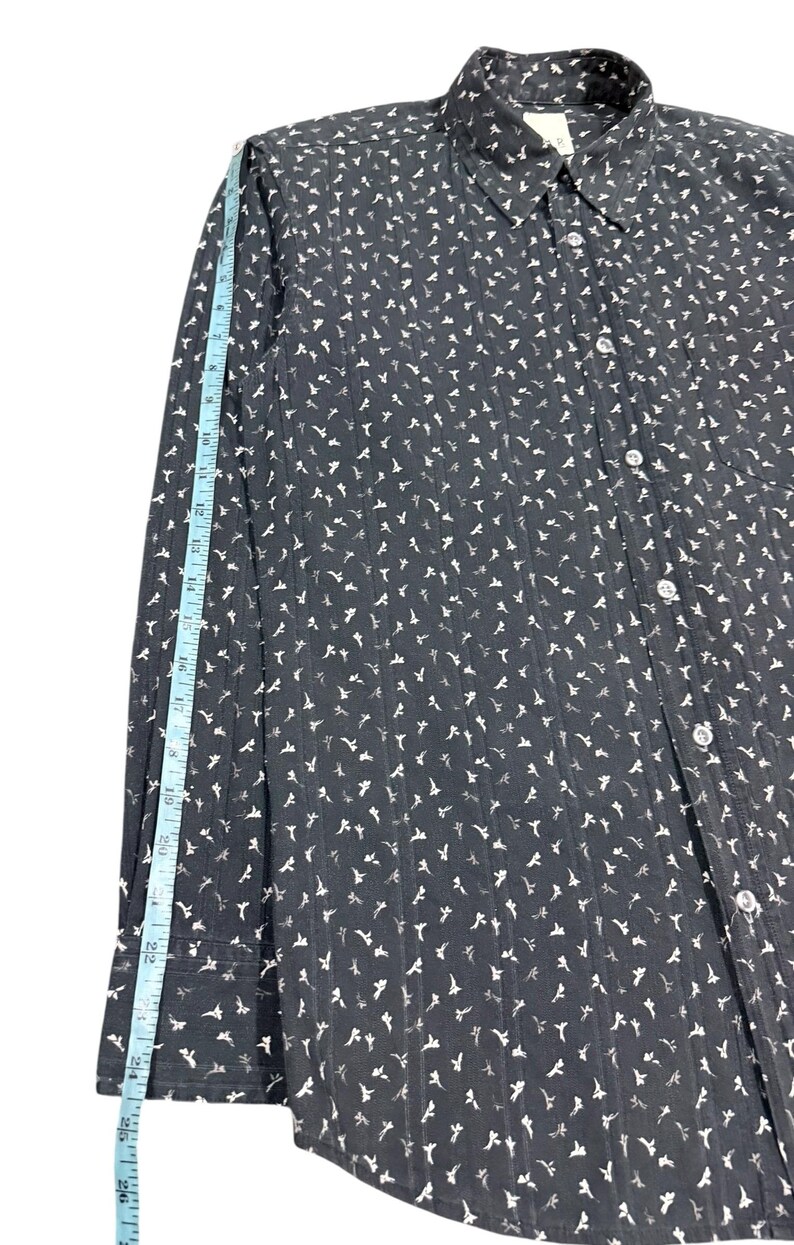 May include: A black long-sleeve button-down shirt with a white floral pattern and vertical textured stripes. A light blue measuring tape is placed along the sleeve, showing a length of approximately 26 inches from the shoulder seam to the cuff.