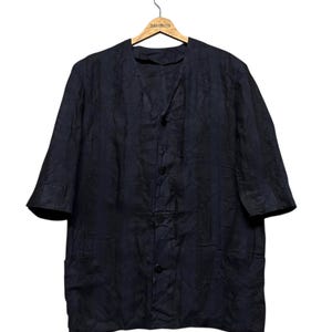 May include: A dark navy blue button-down shirt with a textured, crinkled fabric finish, featuring a collarless V-neckline, elbow-length sleeves, and two front pockets, displayed on a wooden hanger against a plain white background.