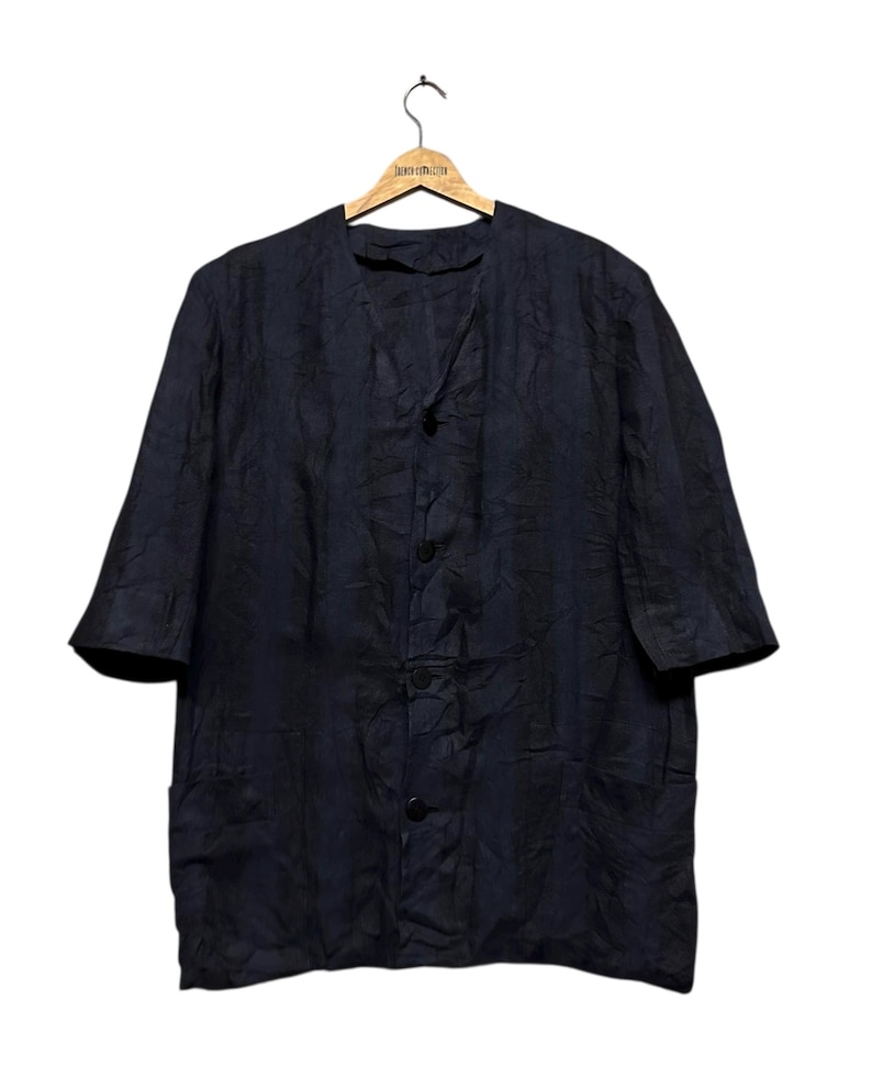 May include: A dark navy blue button-down shirt with a textured, crinkled fabric finish, featuring a collarless V-neckline, elbow-length sleeves, and two front pockets, displayed on a wooden hanger against a plain white background.