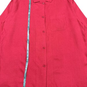 Puede incluir: Una camisa roja brillante con botones, cuello y un bolsillo en el pecho. Una cinta m&eacute;trica azul se coloca verticalmente sobre la camisa. La camisa tiene mangas largas y est&aacute; hecha de una tela texturizada.