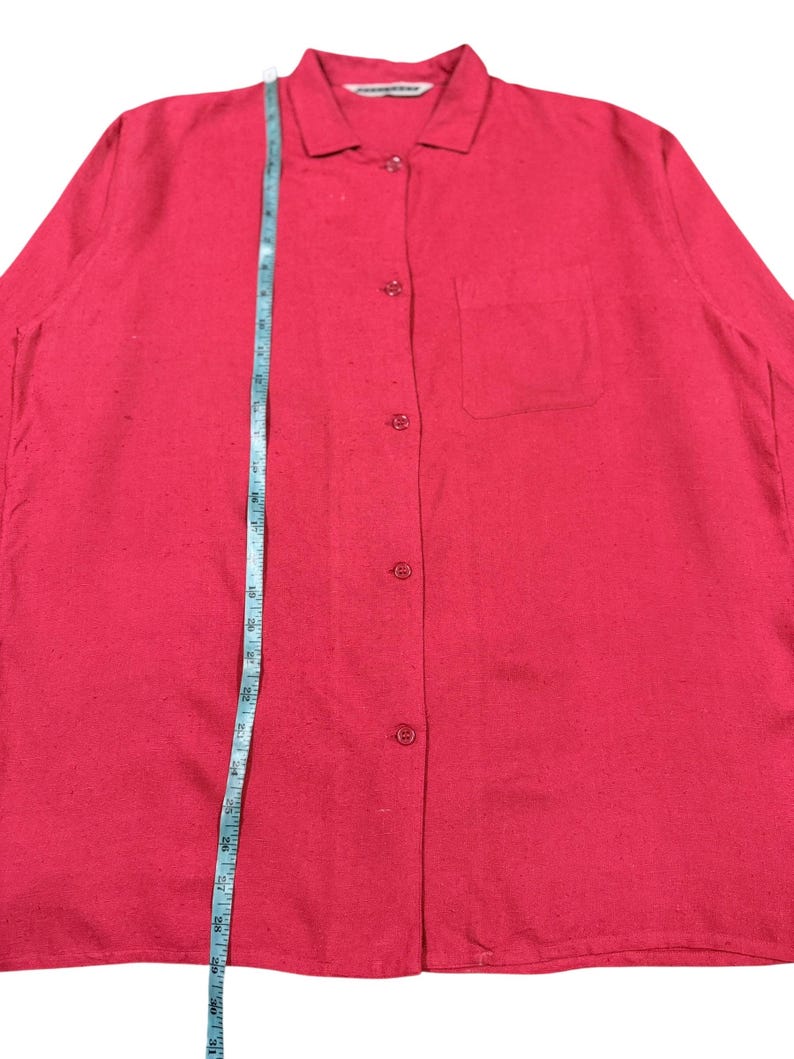 Puede incluir: Una camisa roja brillante con botones, cuello y un bolsillo en el pecho. Una cinta m&eacute;trica azul se coloca verticalmente sobre la camisa. La camisa tiene mangas largas y est&aacute; hecha de una tela texturizada.