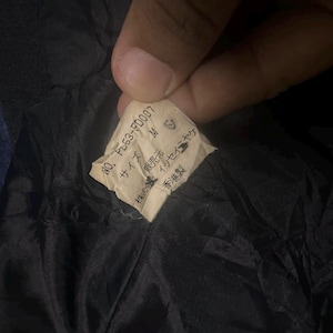 May include: A cream-colored garment care label sewn into a black fabric lining. The label features the text NO. PL53-FD007, size M, and Japanese characters including the brand name Issey Miyake and the manufacturing origin of Hong Kong.