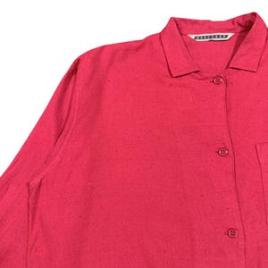 Puede incluir: Primer plano de una camisa roja con cuello, cierre de botones y bolsillo. La camisa tiene mangas largas y una superficie texturizada. El nombre de la marca es visible en el cuello.